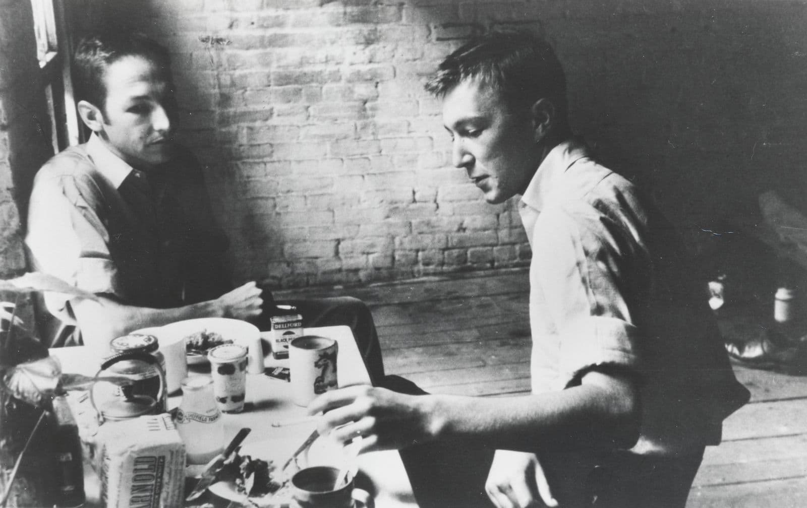Two men sit at a table covered in mugs and art supplies.