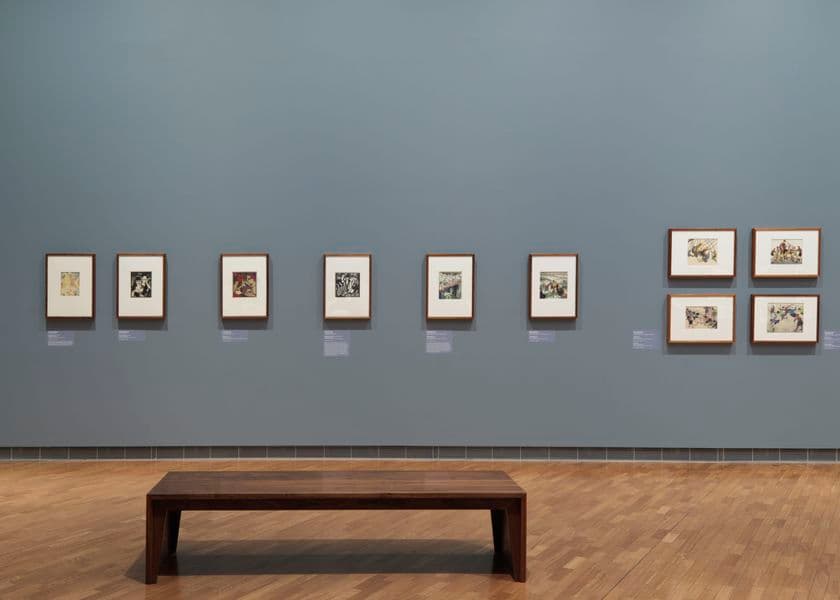 A grey wall with five small works and then a group of four works hanging on the wall