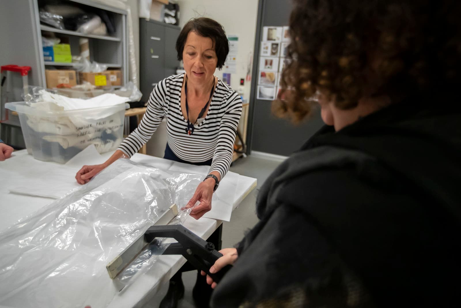 Two women enclose a paper-wrapped object in a plastic bag to undergo preventative conservation