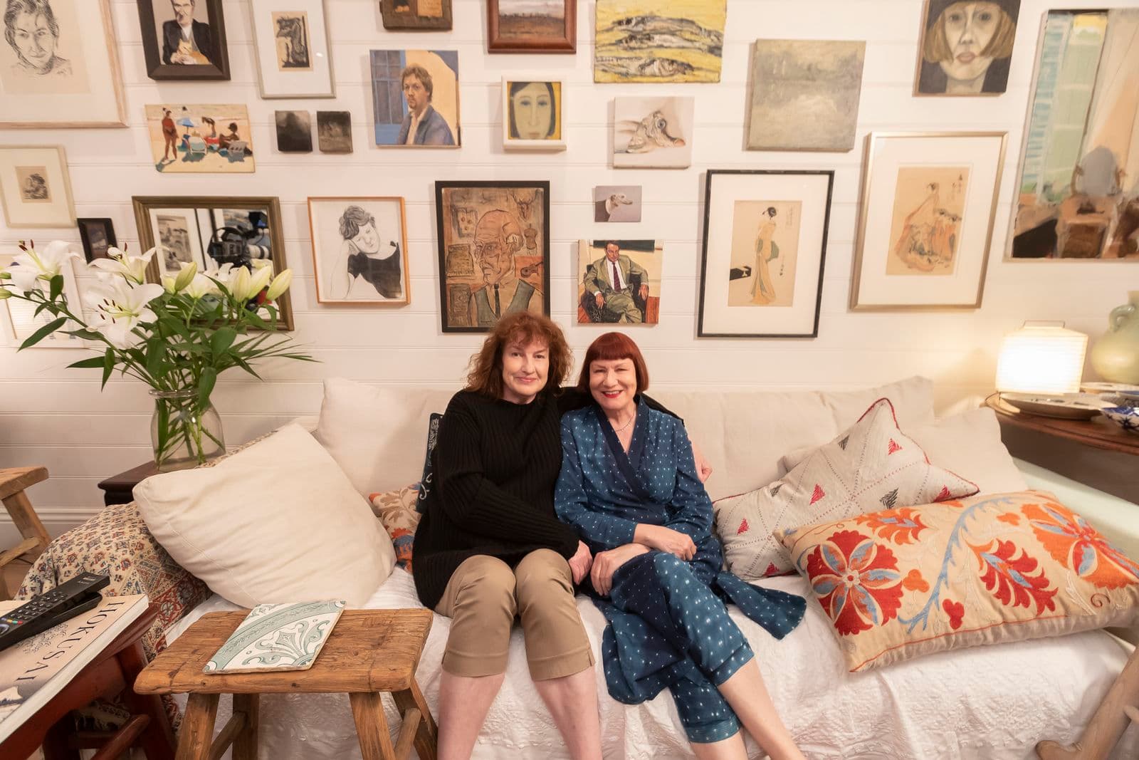 Two smiling women sitting on a couch with a collection of artworks hung on a wall behind them