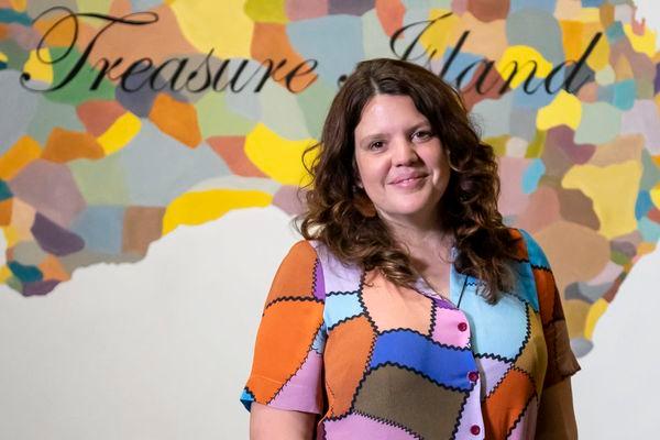 a woman in a bright shirt is smiling and looking at the camera. She is standing in front of a bright painting