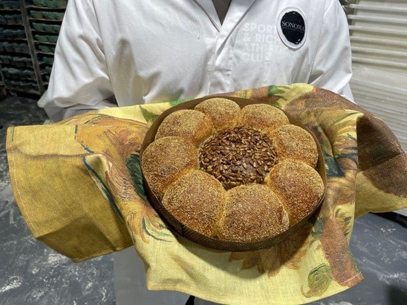 Image of baker holding a circular bread loaf shaped to look like a sunflower with seeds in the middle