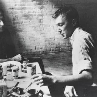 A black and white image of two men sitting around a rectangular table with crockery and food, and a brick wall behind them
