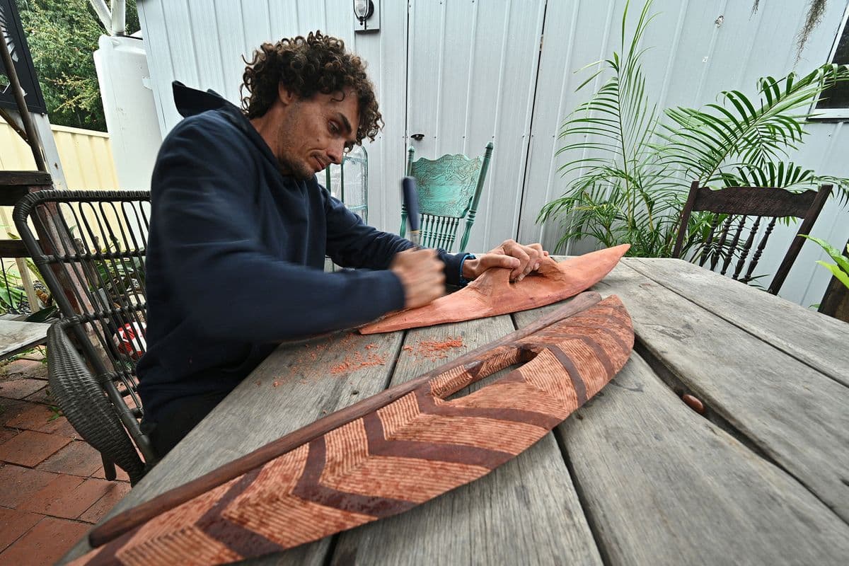 A man sits at a table carving a wooden shield