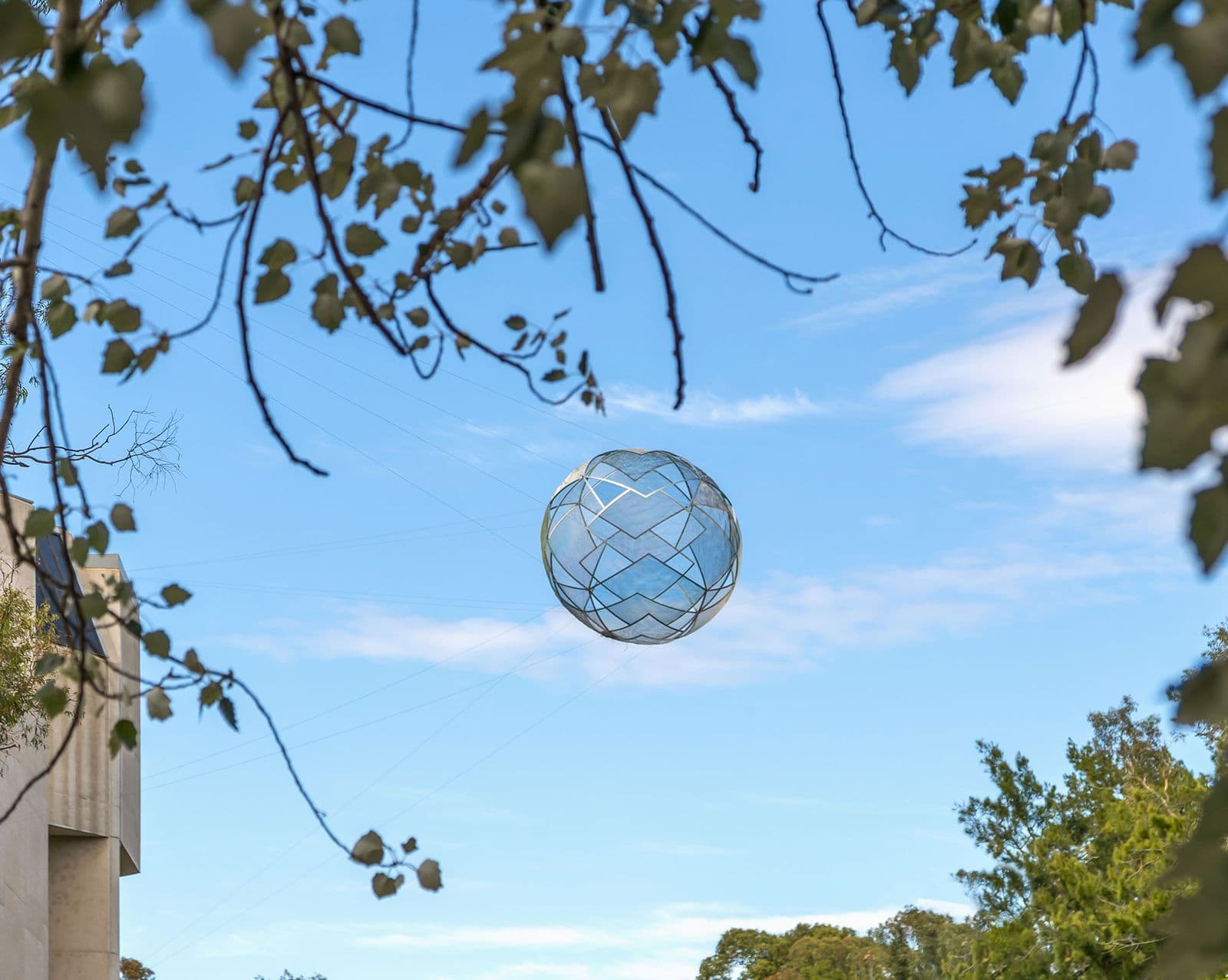 A large spherical sculpture hanging from large wires in the sky is visible through a gap in tree branches