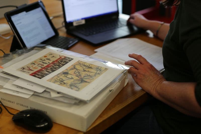 Photograph of woman seated at table with laptop and archival material surrounding her