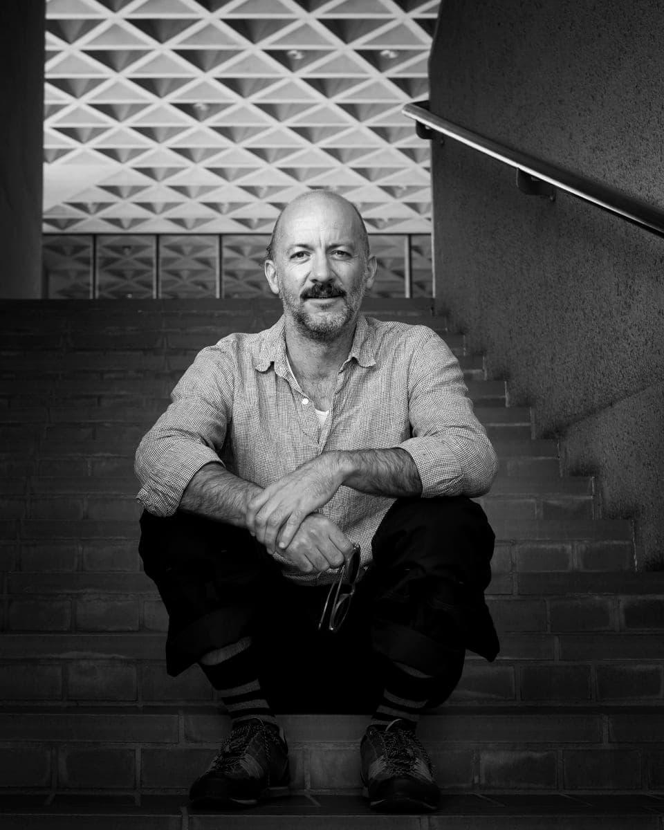 Portrait of a white male, blue shirt, black pants seated on steps with the Col Madigan triangular featured architectural roof