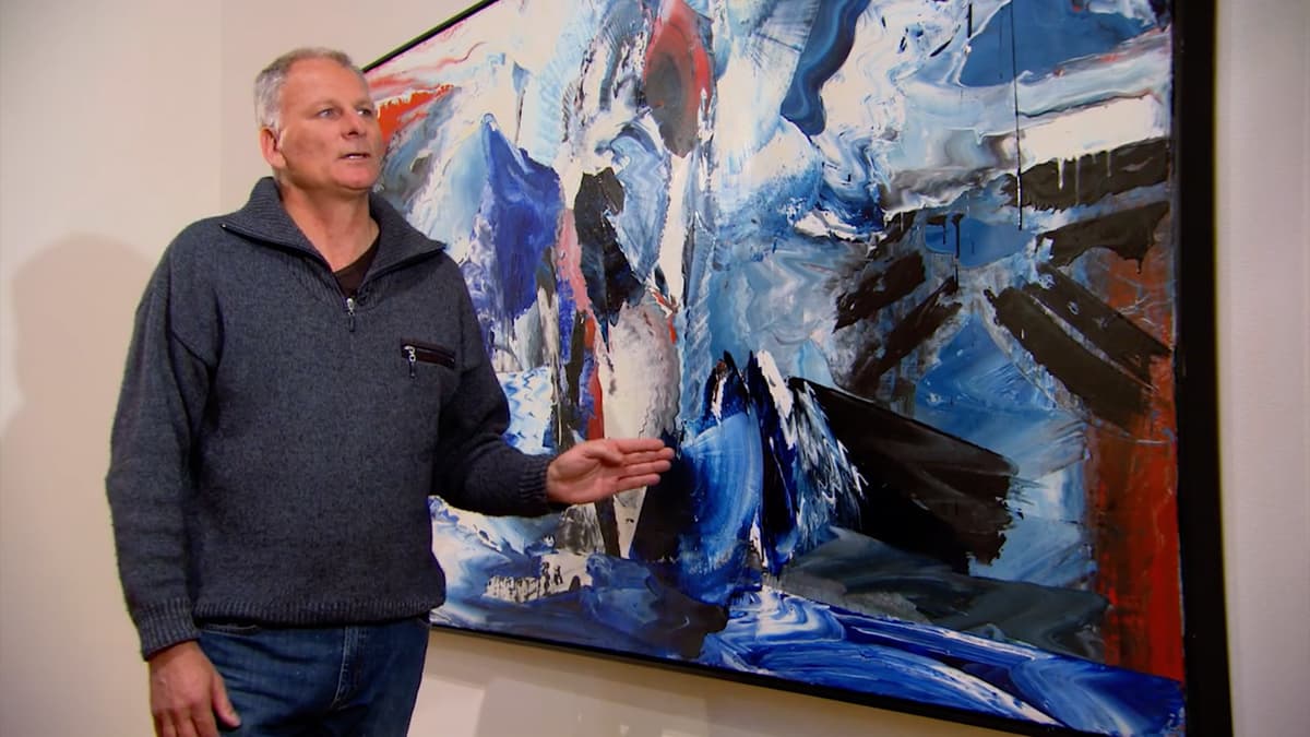 A video still of a man speaking to the camera in front of a large painting