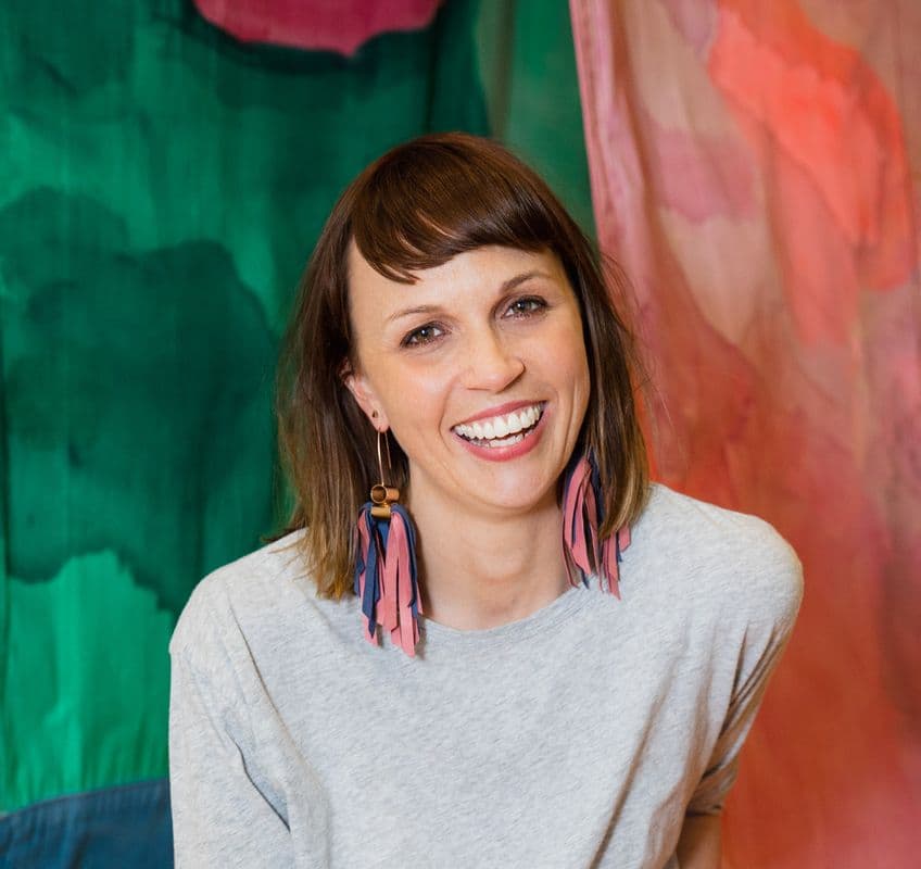 Portrait of female smiling, with mid length brown hair wearing colour tassel earrings, in front of a colourful background
