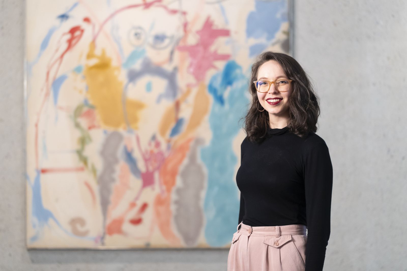 A woman with glasses standing in front of an abstract painting in pastel tones.