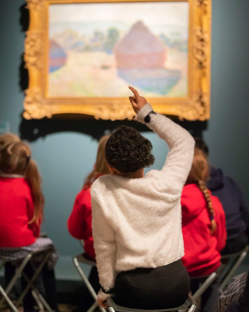 Photograph of school children sitting in front of painting with arms raised to answer question