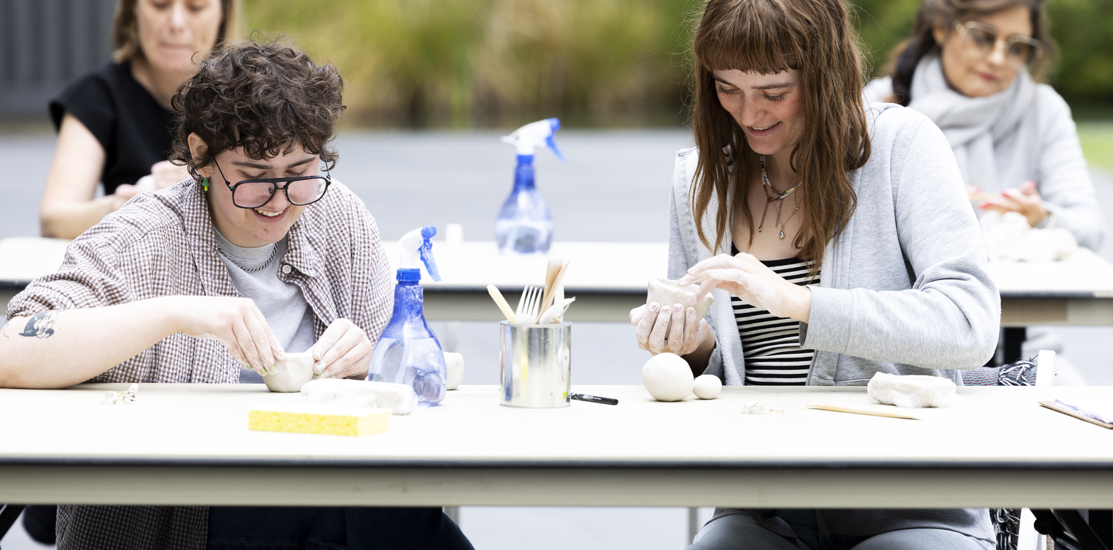 Photo of two people working on clay over a table outdoors