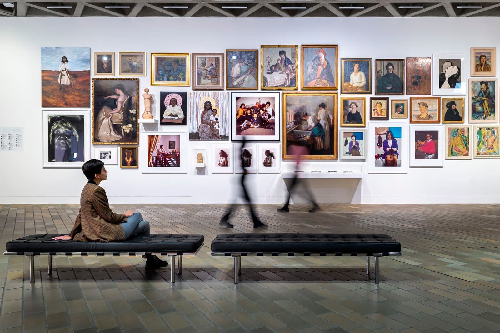 A woman is sitting in font of a large wall of many portraits in a large gallery space