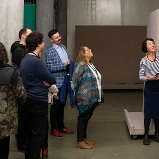 Five people are watching a demonstration given by a woman in a painting storage area.