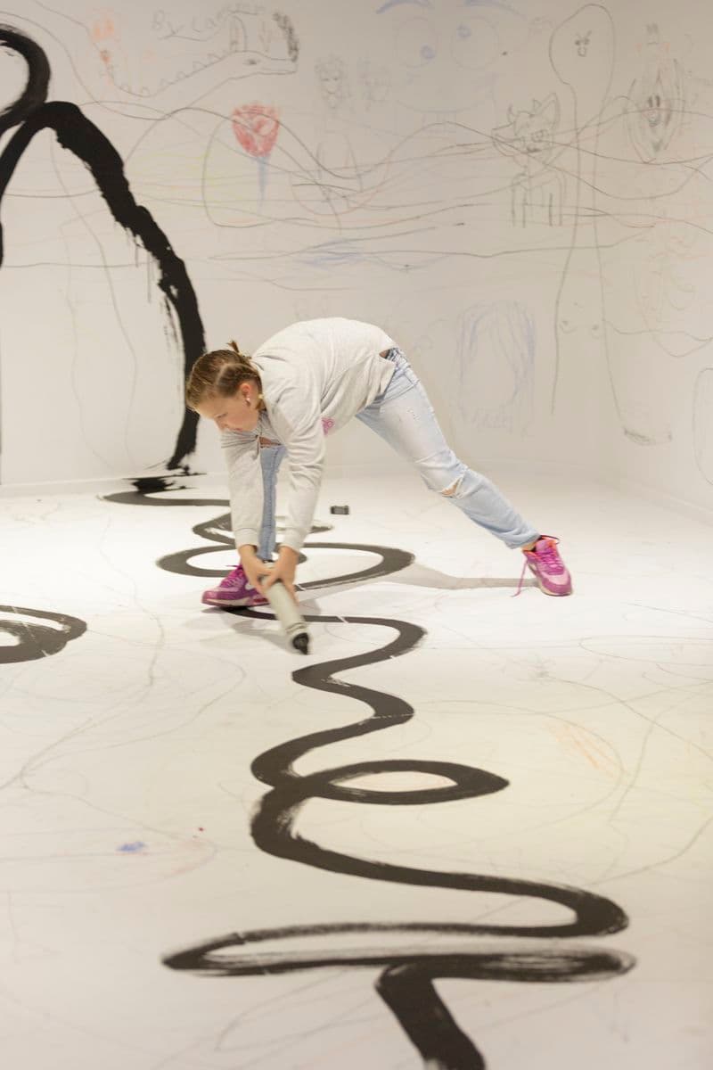 In a room with drawing marks and scribbles all over the white floor and walls child is drawing on the floor using an oversize crayon
