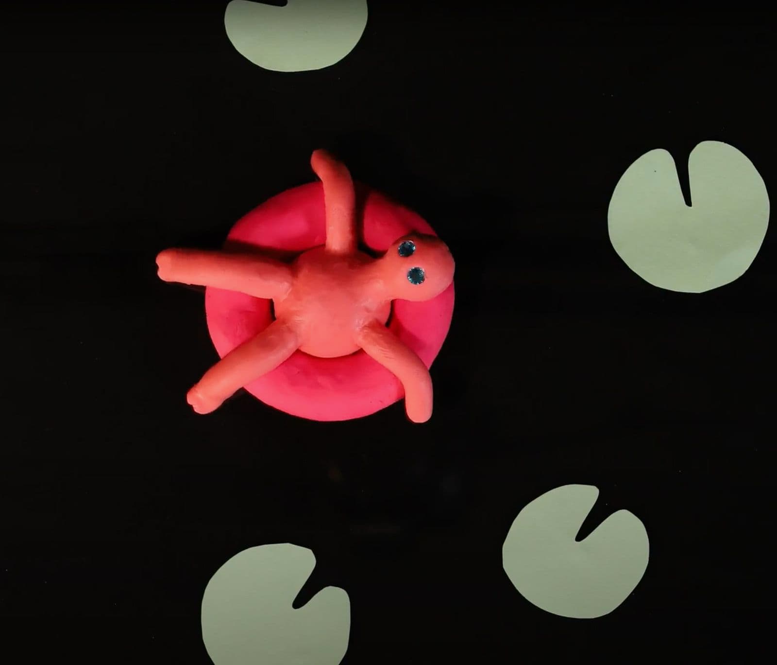 Screenshot of a stop motion video of a black background and a red plasticine shaped person lying in a red lilo surrounded by green plasticine shaped lillypads.