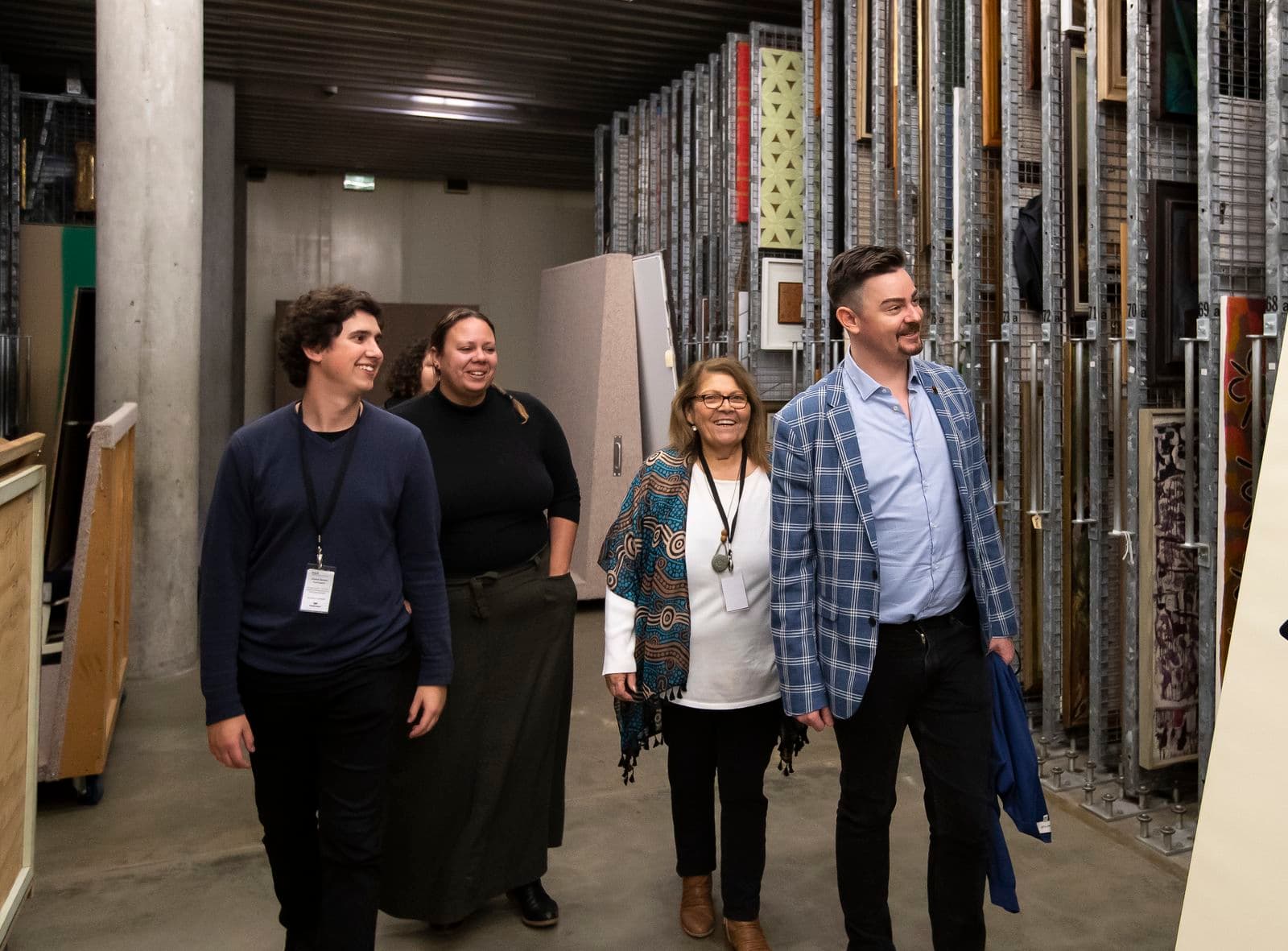 Four people are walking in a large painting storage room, with many paintings hanging in tall wire storage racks
