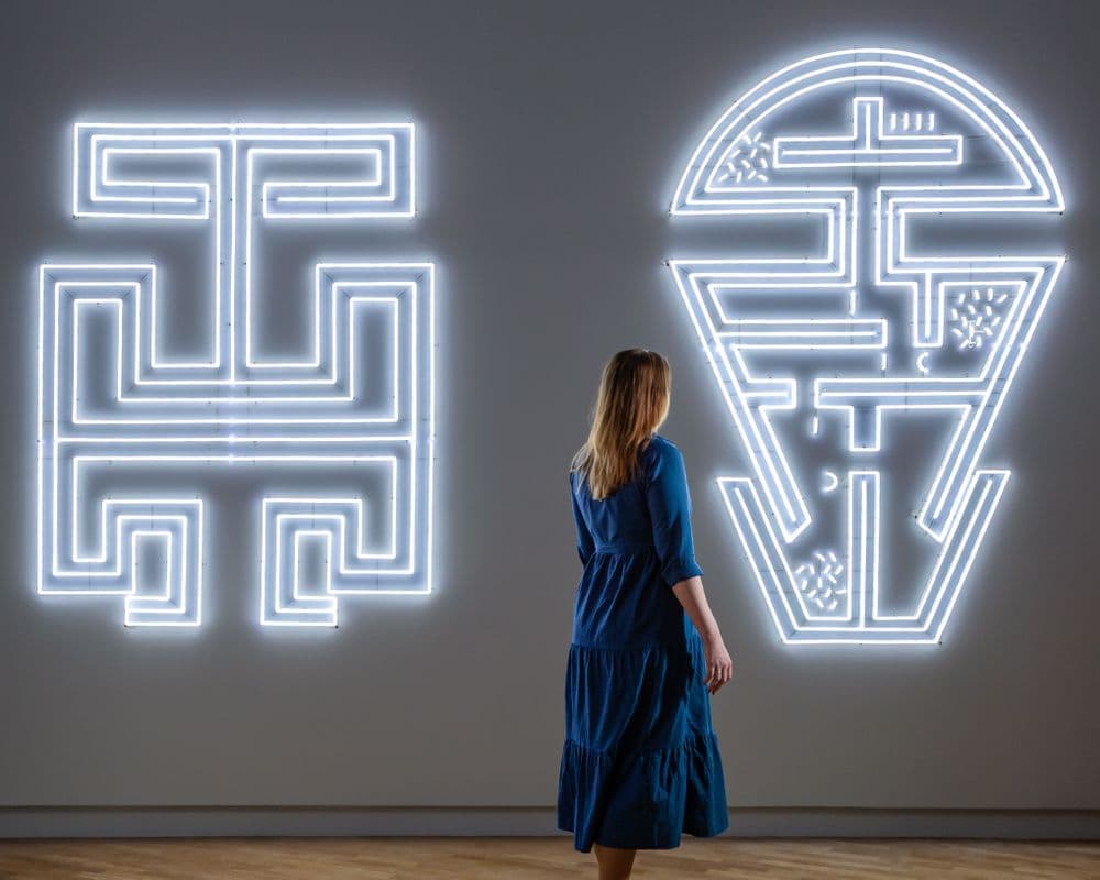 A woman standing in front of two large linework neon light designs are hung across a wide wall