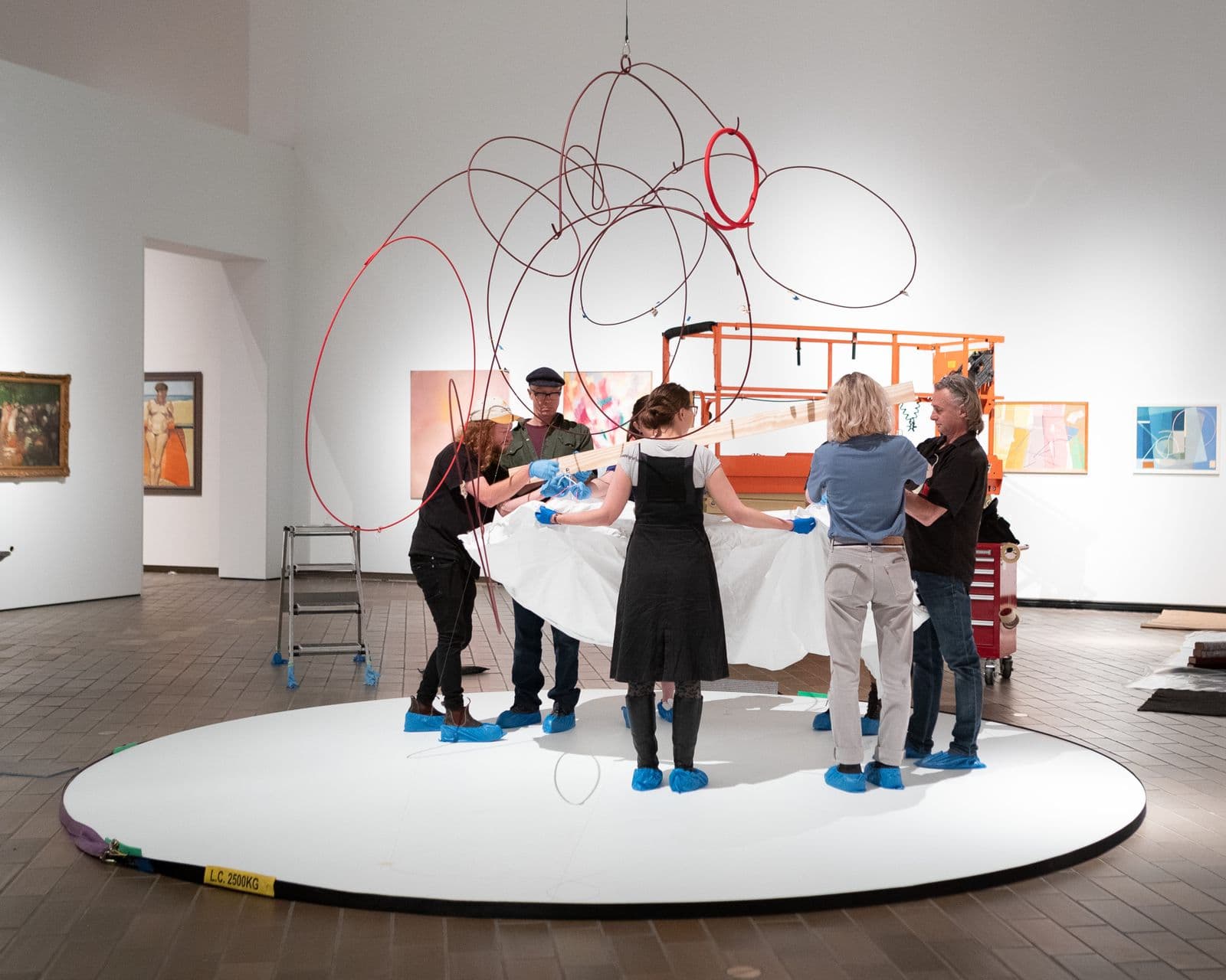 A group of five people in the centre of a gallery space hold a large sheet as they install a large hanging mobile from the ceiling