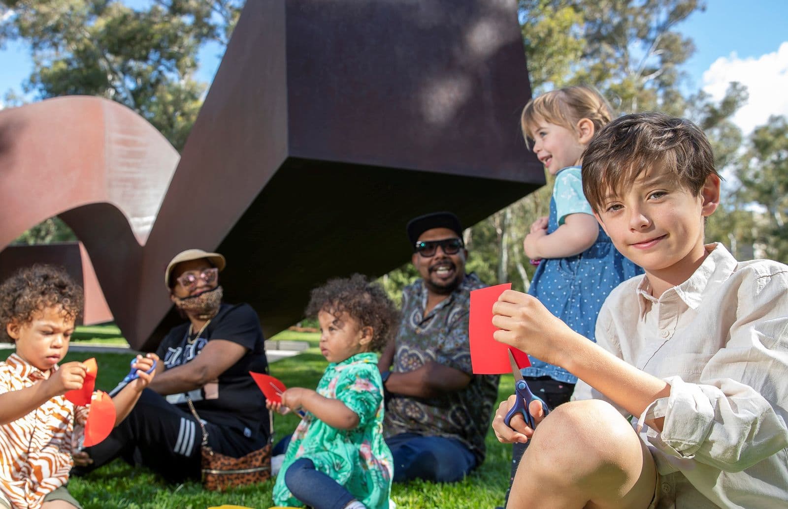 Two adults sit with four children in front of a sculpture in the sculpture garden making art with paper