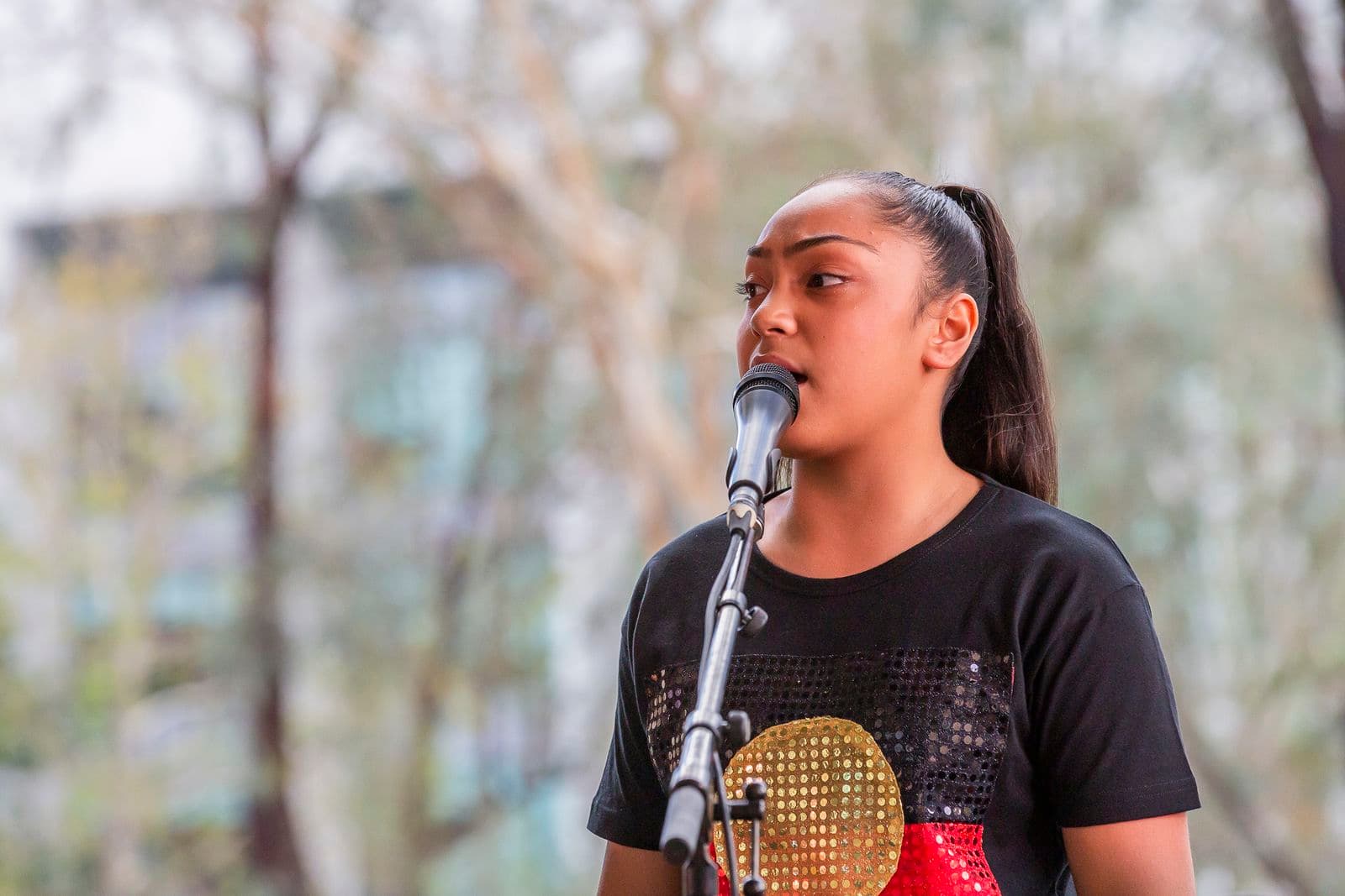 Photograph of a young person wearing a t-shirt with the Indigenous flag singing into a microphone