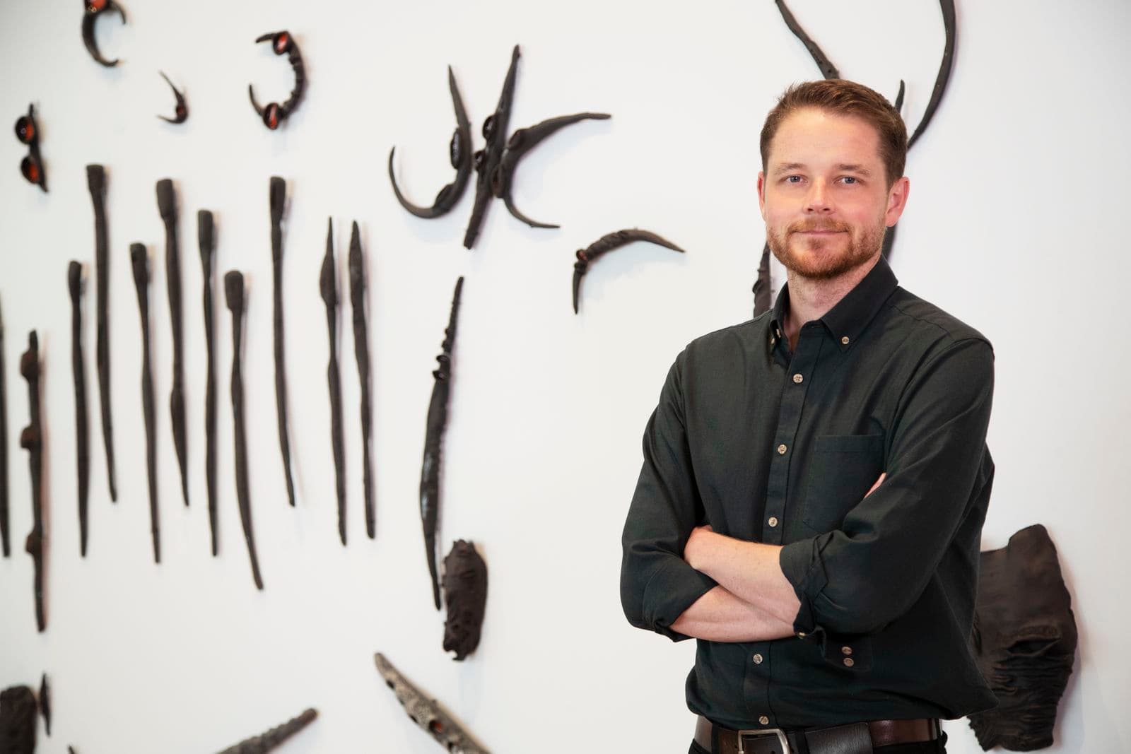 A man stands with his arms crossed in front of a white wall with black ceramic banksia shapes.