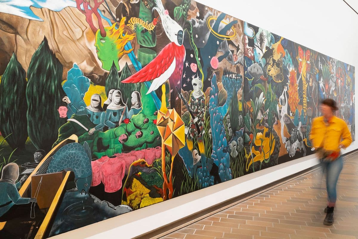 A woman in a yellow jumper appears blurred while walking by a large painting hung in a gallery