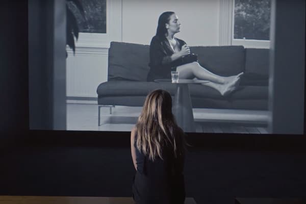 Screen shot of a film still showing a woman from behind, sitting on a bench and watching a large film projection