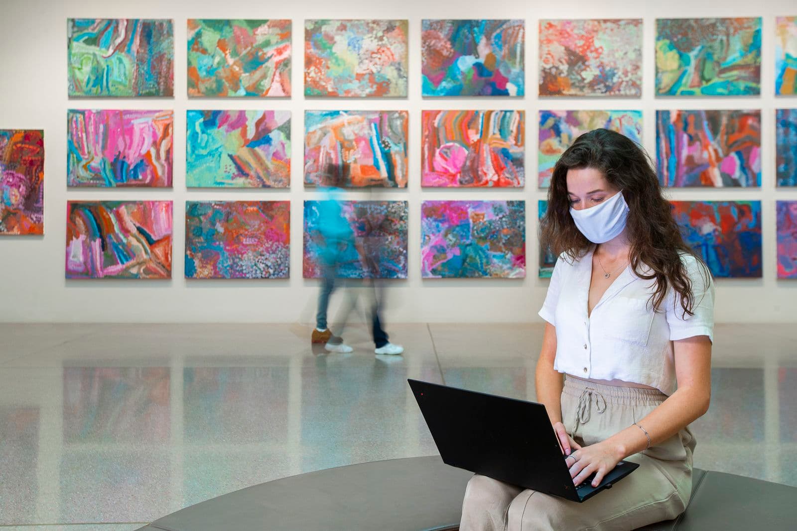 Young woman sitting in a Gallery space wearing a face mask, typing on laptop