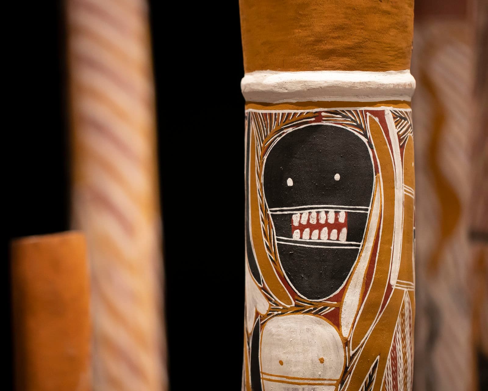 A close-up photograph showing a painted hollow-log. It is part of an installation of 200 painted hollow logs standing upright in a large gallery space. The surrounding walls are painted in a dark colour.