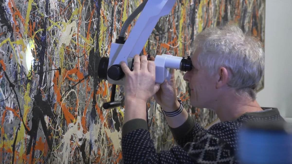 Video still of man looking at painting closely through a microscope.