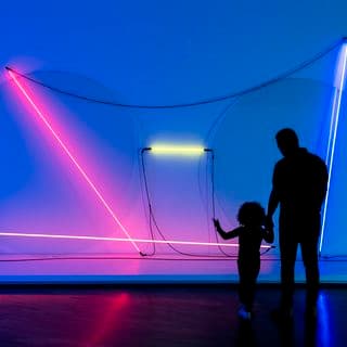 An adult and a child are silhouetted as they stand in front of a bright light installation made of neon tubing affixed to a wall