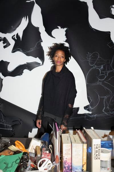 A portrait of the artist Kara Walker - an African American woman standing against a black and white background of her painting. Books and art making materials in the foreground
