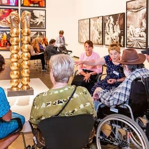 a group of people, some in wheelchairs, sit around a sculpture listening to a guide