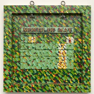 A sculpture of a timber frame fashioned as a calendar month with large text reading "Month of May" and small squares underneath acting as calendar days with numbers, covered in green, yellow and red paintbrush strokes