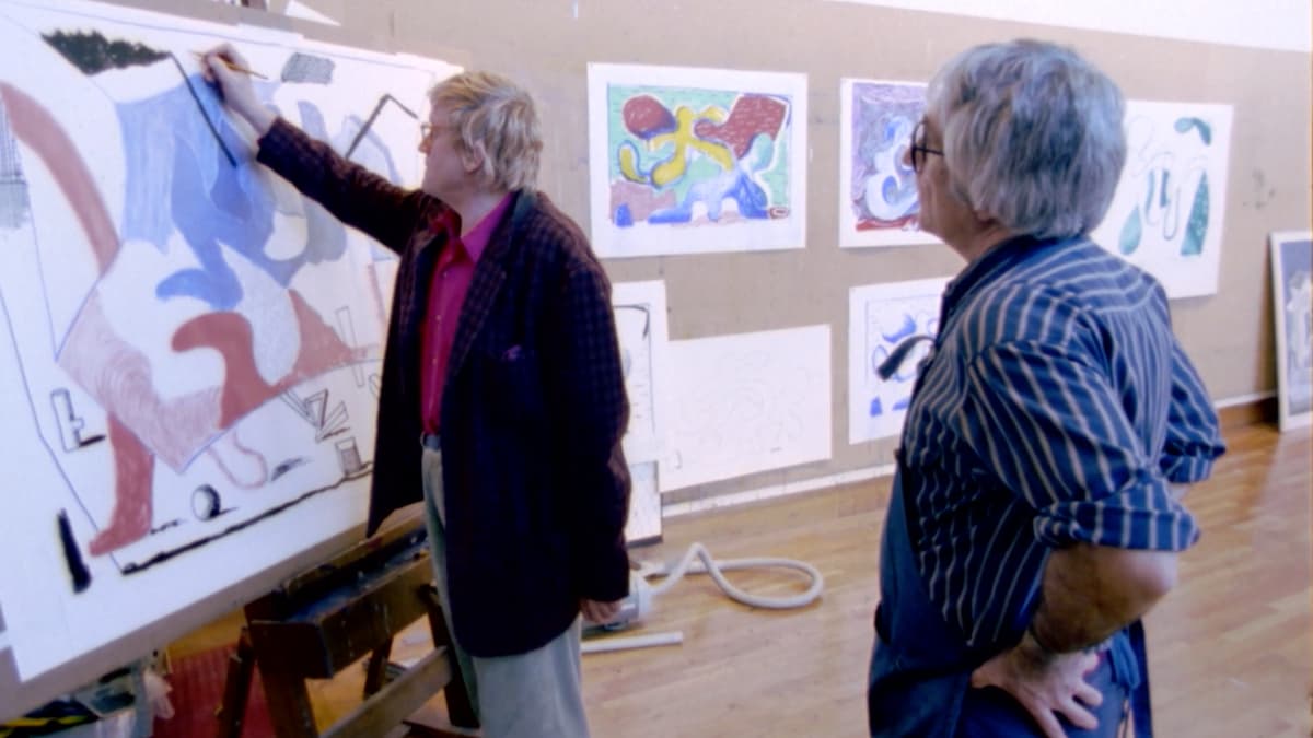 A video still of two men in a studio, one drawing and another onlooking