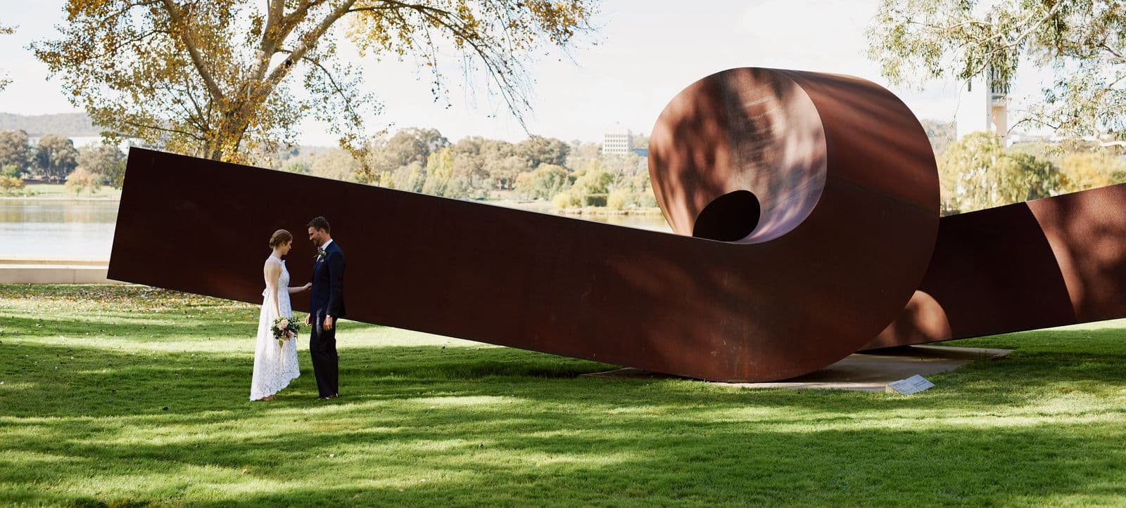 A couple stand, facing each other and holding hands, in front of a large sculpture in a garden
