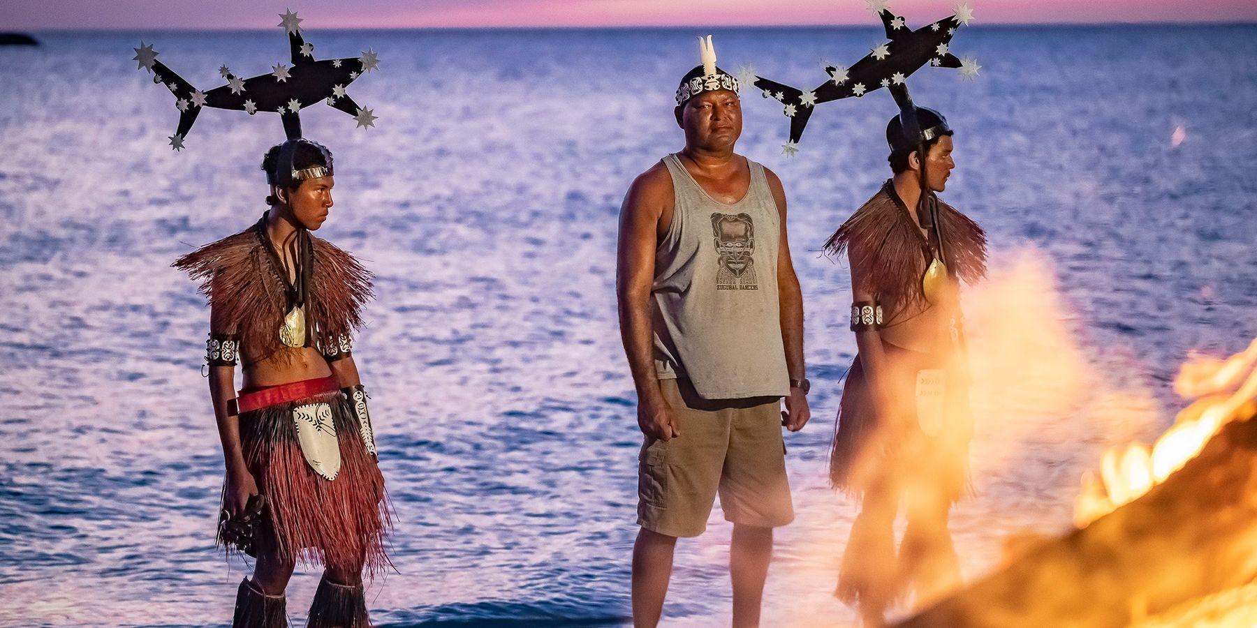 Photograph of artist Alick Tipoti standing on a beach on Badhu Island, in the Zenadth Kes/Torres Straits at sunset with other members of his community is ceremony dresss