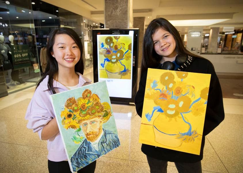 Two children stand in a shopping mall holding paintings they have done representing Van Gogh's Sunflowers