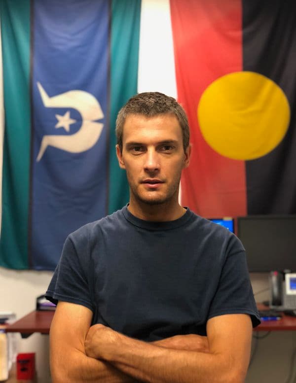 Portrait of young male with short cropped hair, wearing a dark t-shirt with his arms folded, standing in front of the Torres Strait Islander Flag and Aboriginal Flag