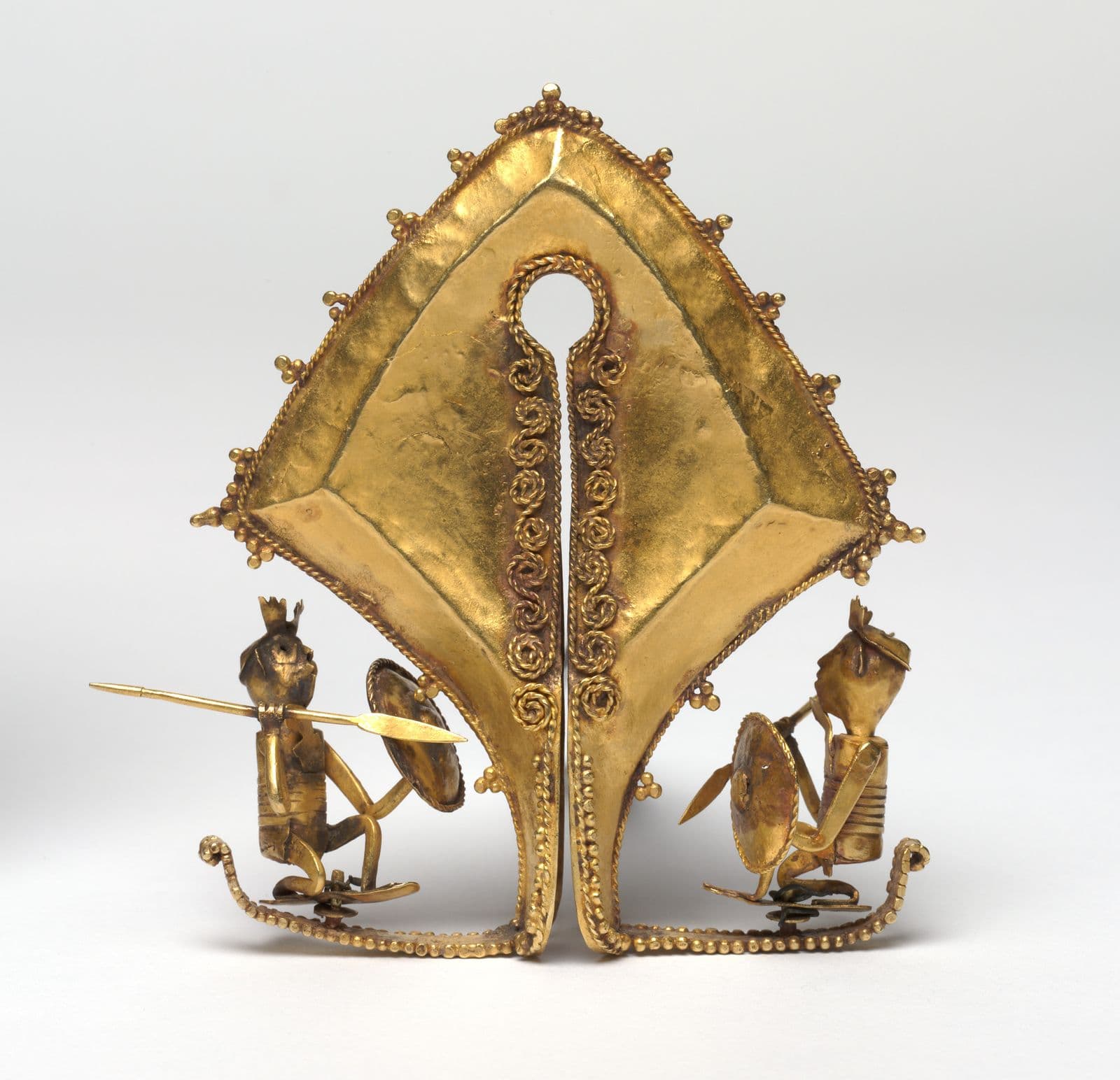 Gold ceremonial ear pendant, two warrior figures wielding spears and shields lie on either side of a diamond shaped central shape.