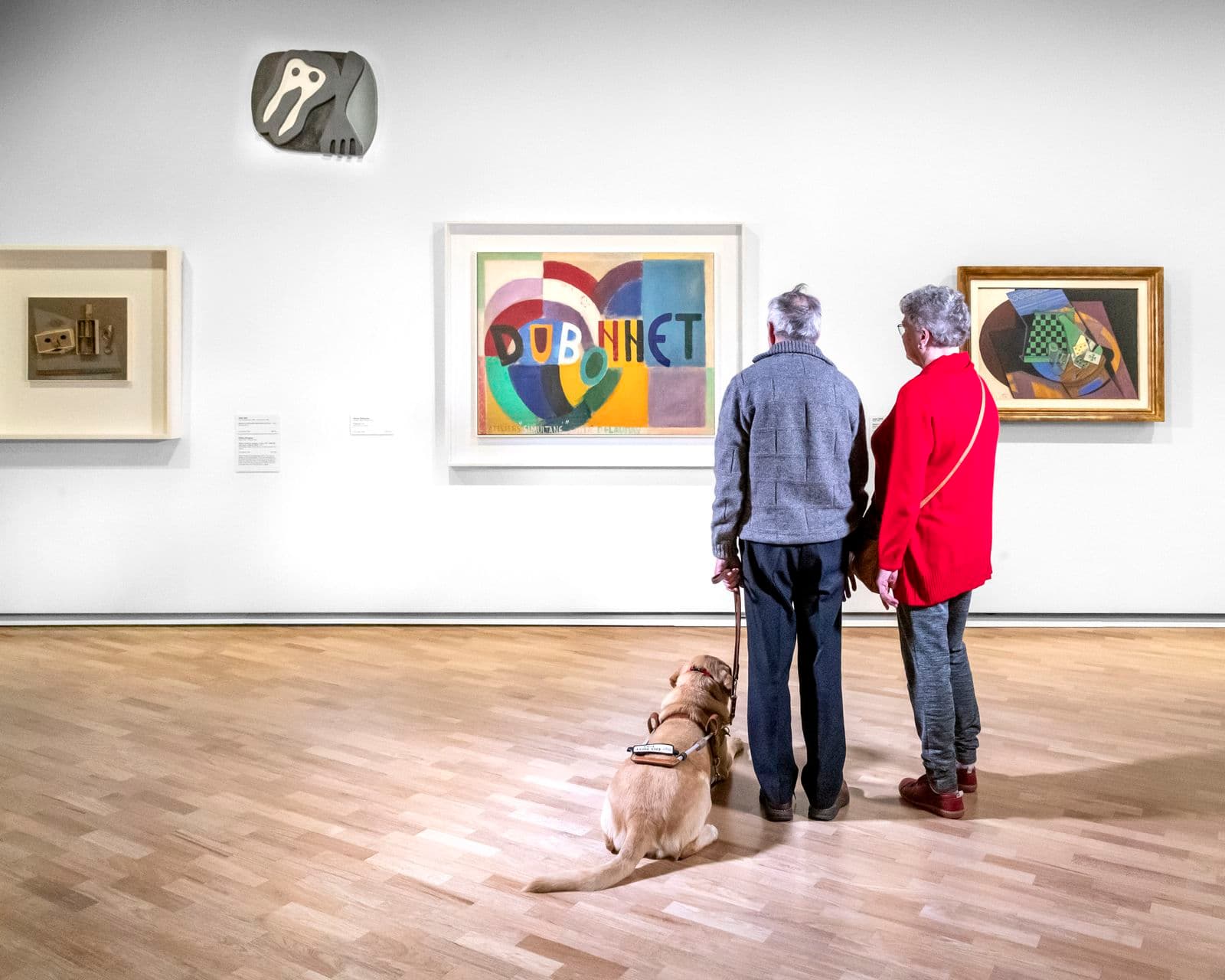 Photograph of two visitors facing artworks with companion dog by side