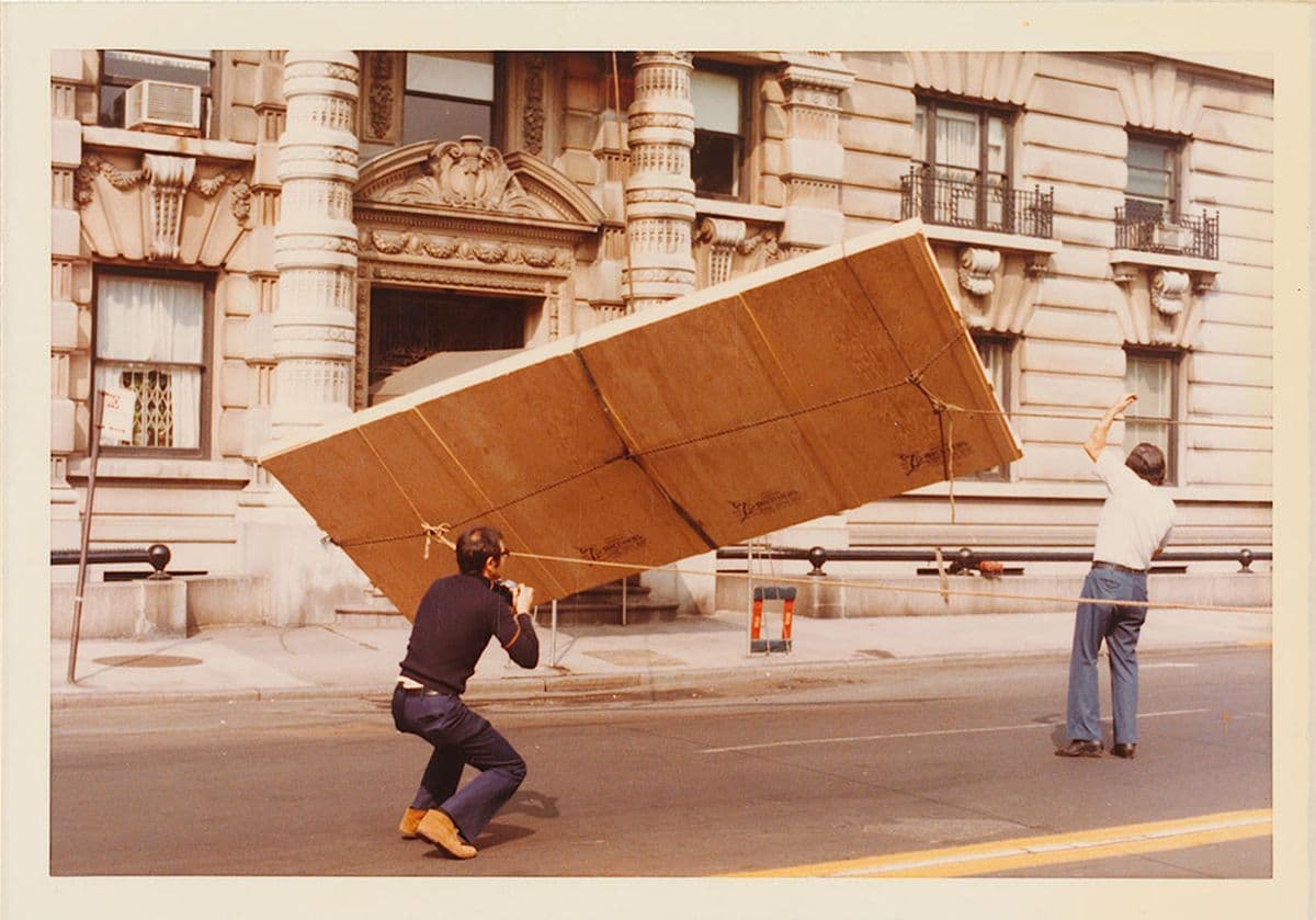 Two men using a pulley system to lower a boxed painting from a tall apartment block