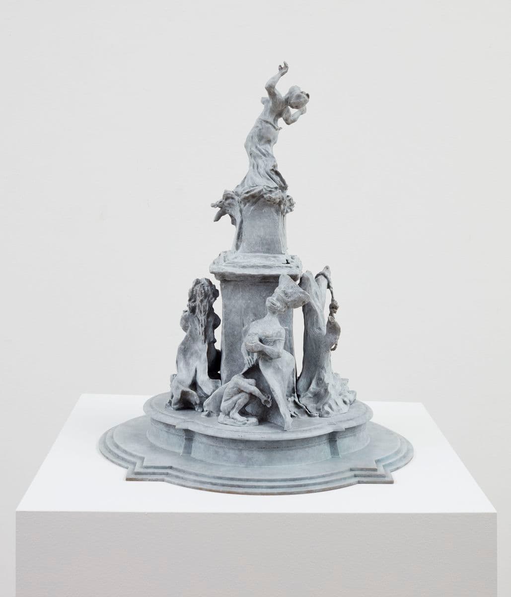 An image of a sculpture on a plinth with a grey fountain structure and multiple figures