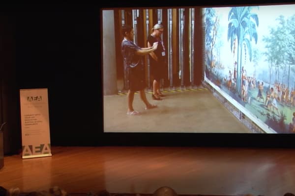 A video still of a woman giving a lecture with a projection to the right