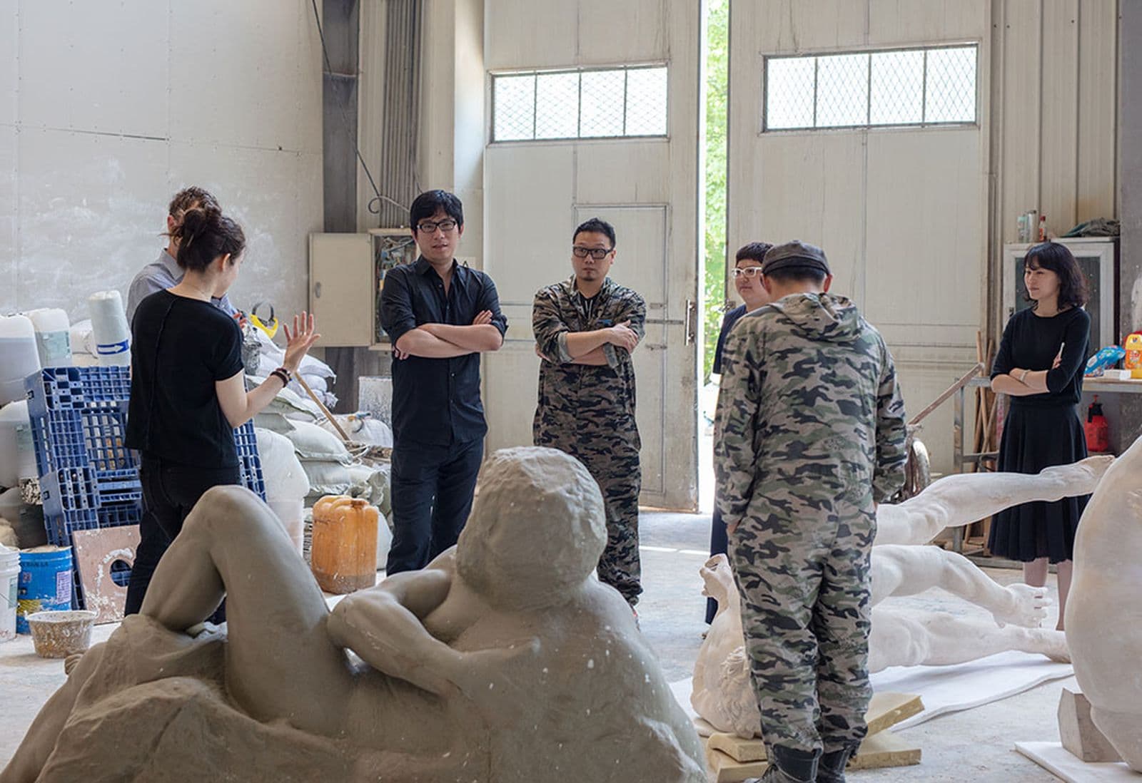 A group of people stand in a circle in an artists studio filled with sculptures