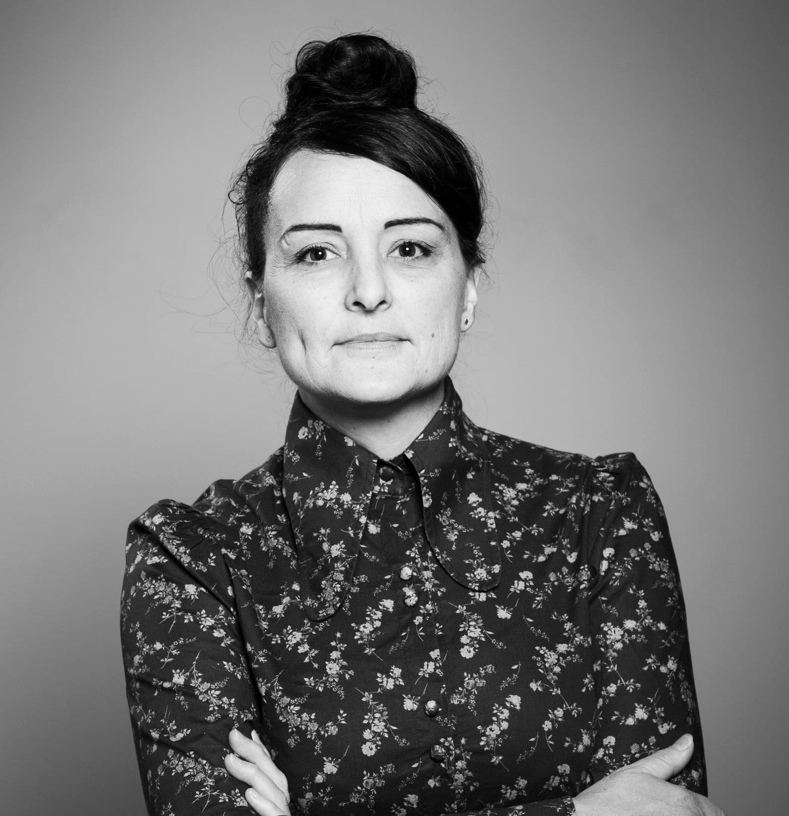 Black and white photo of  woman standing, wearing a buttoned up floral shirt, with her hair up in a bun.