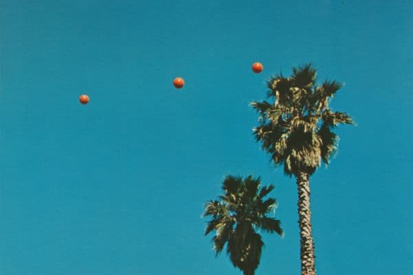 Three orange balls are mid-air with a bright blue sky and the tops of two palm trees in the background