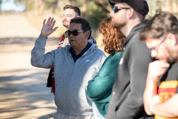 A man wearing a grey zip-up jumper is gesticulating with his arm while speaking to a group of people standing on either side of him