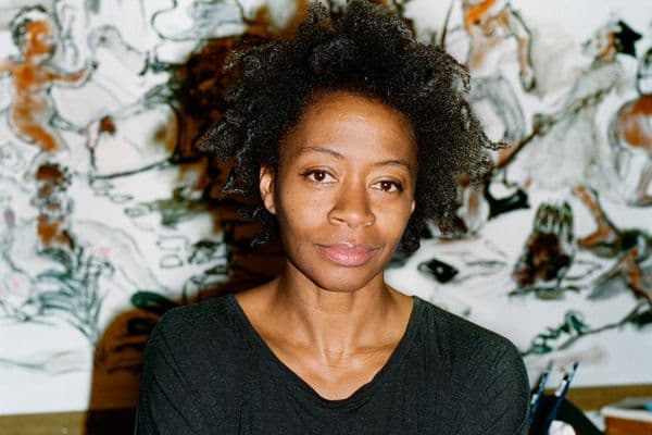 A photographic portrait of a woman artist in a black t-shirt with studio works in the background